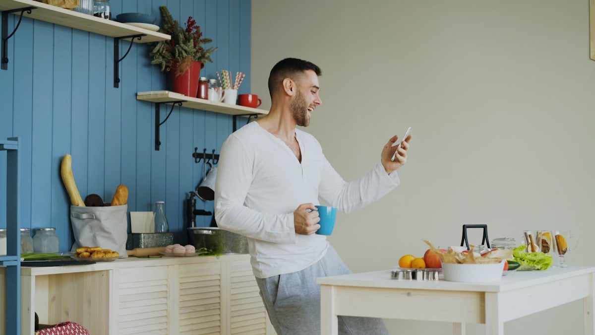 Cheerful happy man dancing and singing in kitchen while browsing social media on his smartphone at home in the morning