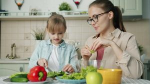Cute loving daughter is helping mother with salad cutting cucumbers feeding young woman and laughing having fun at home. People and cookery concept.